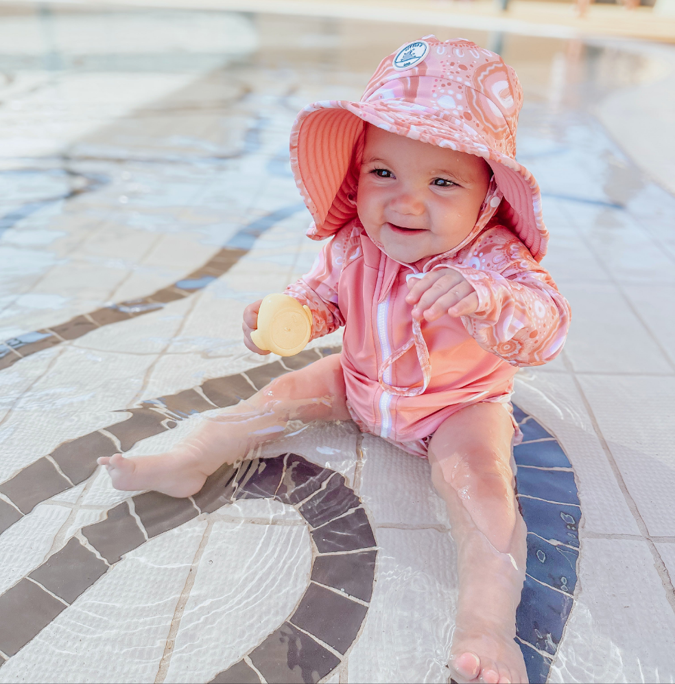 LITTLE E & CO - four-piece kids' swim set featuring a long-sleeve rash guard, swim hat, swim shorts and wet bag. The set is a peach pink with a colourful contemporary indigenous print. Made with UPF 50+ sun protection fabric. The rash guard has a front zipper for easy wear, and the shorts feature an adjustable drawstring waistband for a secure fit, and the hat has adjustable toggles.