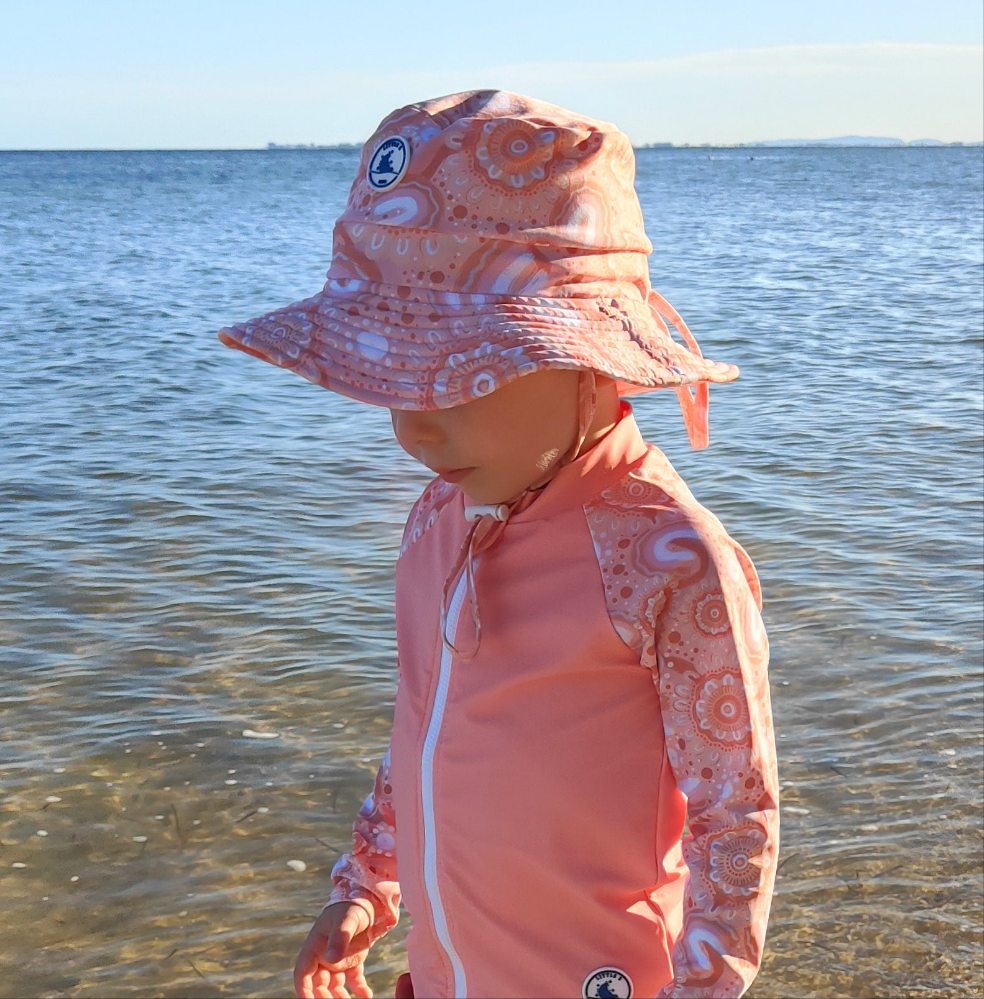 Child wearing a pink sun hat and matching outfit of kids rashie by the water