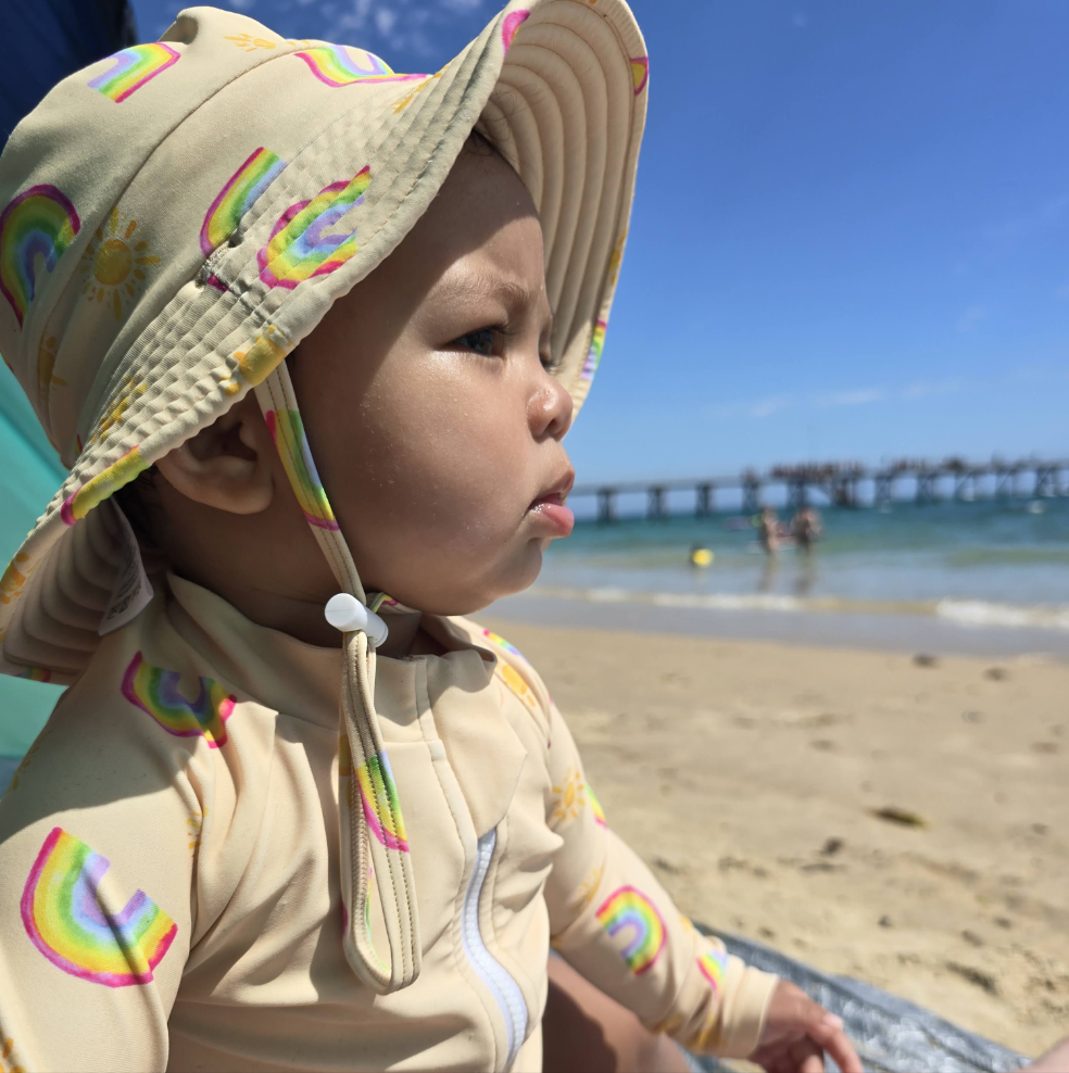 Baby swimwear set in pale yellow with rainbow and sun print, featuring long sleeve rashie, adjustable swim nappy, and sun hat, worn by toddler enjoying beach view.