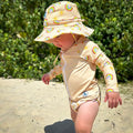 Baby swimwear set with pale yellow rash guard, swim nappy, and hat in rainbow and sun print, shown on child outdoors, designed with UPF 50+ sun protection.
