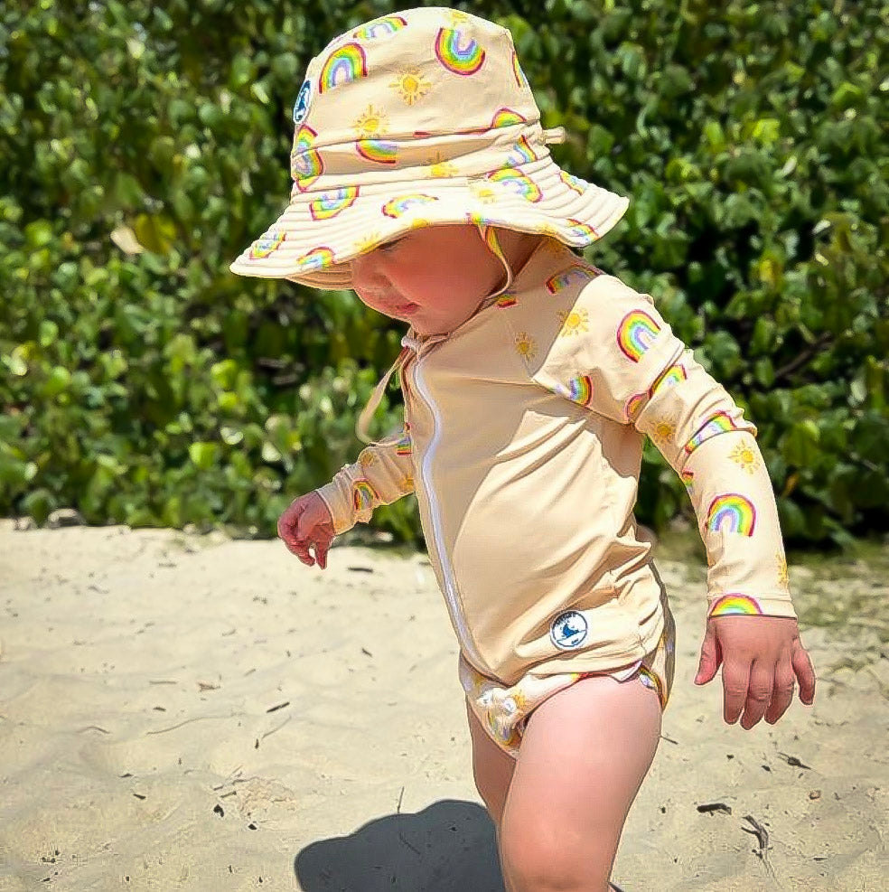 Baby swimwear set with pale yellow rash guard, swim nappy, and hat in rainbow and sun print, shown on child outdoors, designed with UPF 50+ sun protection.