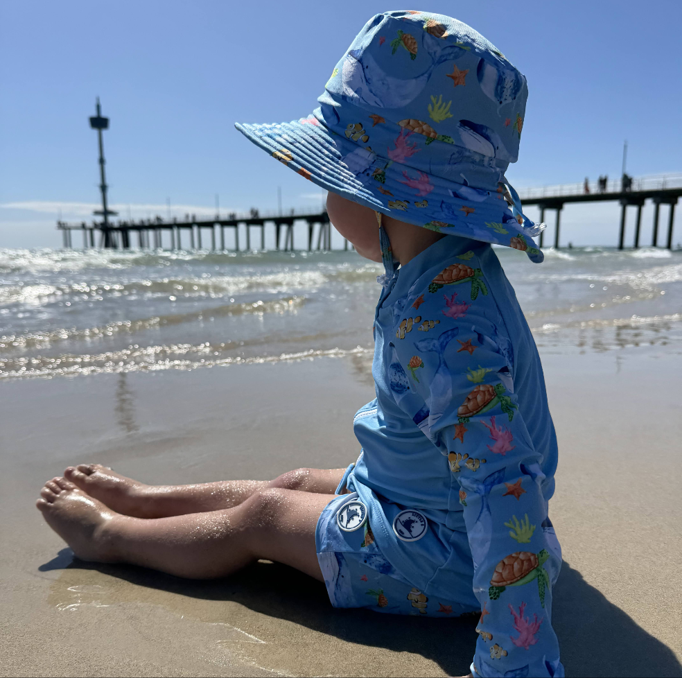 LITTLE E & CO - four-piece kids' swim set featuring a long-sleeve rash guard, swim hat, swim shorts and wet bag. The set is blue with a colourful ocean print featuring whales, turtles, fish and coral. Made with UPF 50+ sun protection fabric. The rash guard has a front zipper for easy wear, and the shorts feature an adjustable drawstring waistband for a secure fit, and the hat has adjustable toggles.