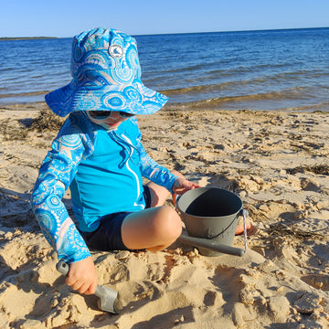 Baby swimwear outfit in turquoise blue with vibrant Indigenous print, including long-sleeve rashie, adjustable swim nappy, sun hat, and matching wet bag.