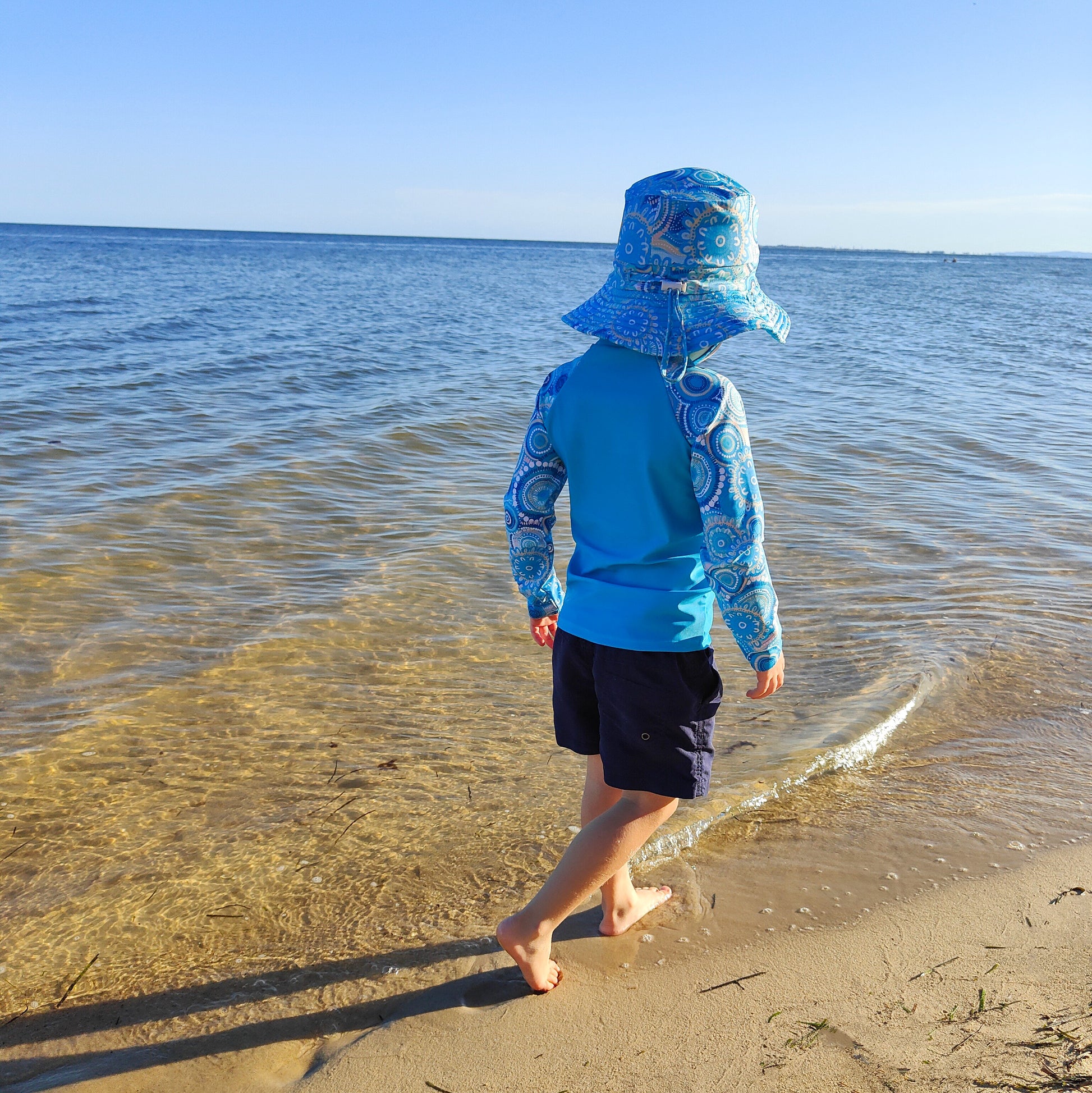Baby swimwear set in turquoise blue with contemporary Indigenous print, featuring a kids’ long-sleeve rash guard and sun hat with adjustable toggles.