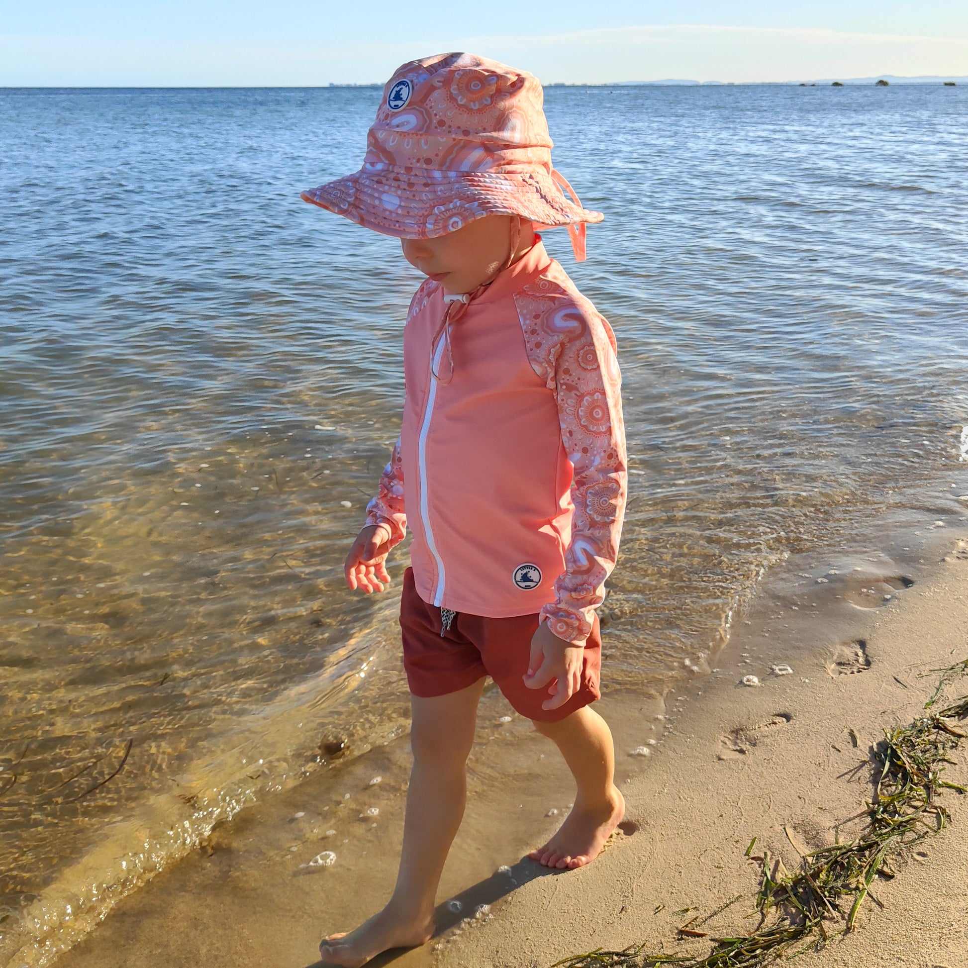Baby Swimwear Set in peach pink with contemporary indigenous print, includes UPF 50+ kids long sleeve rashie with front zipper and sun hat with adjustable toggles.