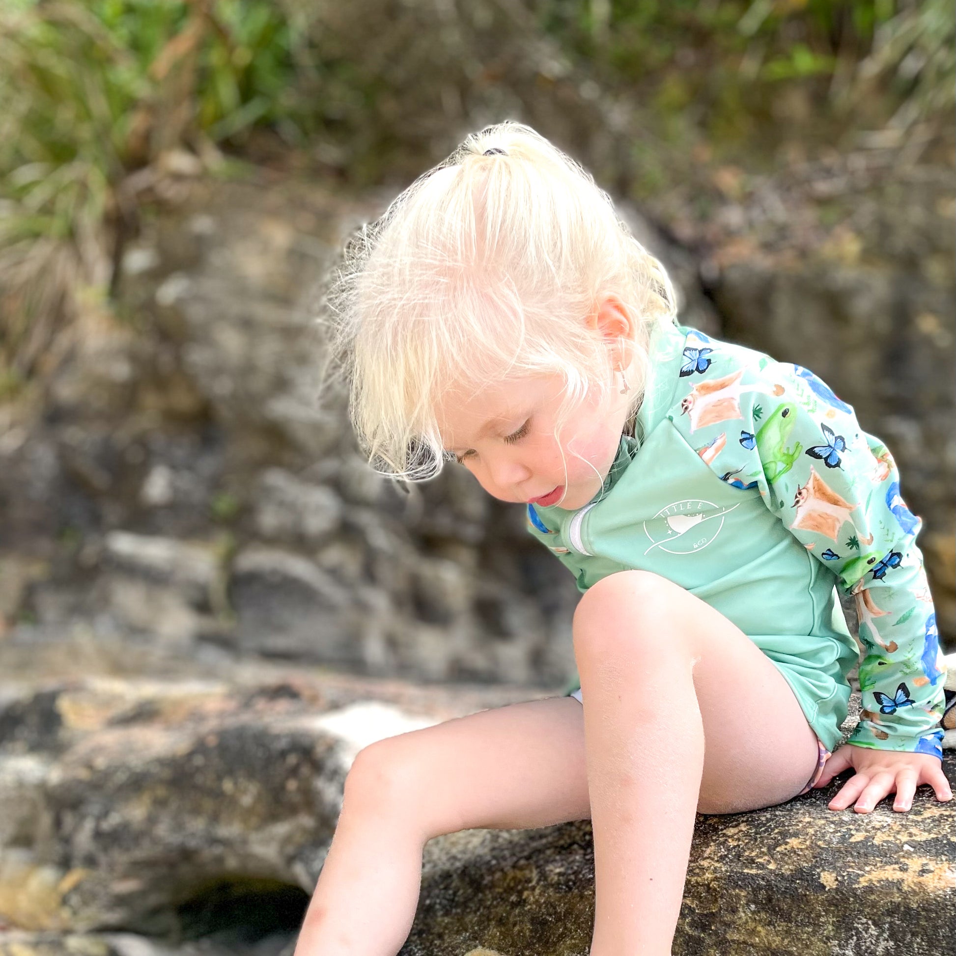LITTLE E & CO – kids long-sleeve rash guard, a pastel purple with a green with a colourful rainforest print featuring birds, frogs, lizards and butterflies. Made with UPF 50+ sun protection fabric. The rash guard has a front zipper for easy wear.