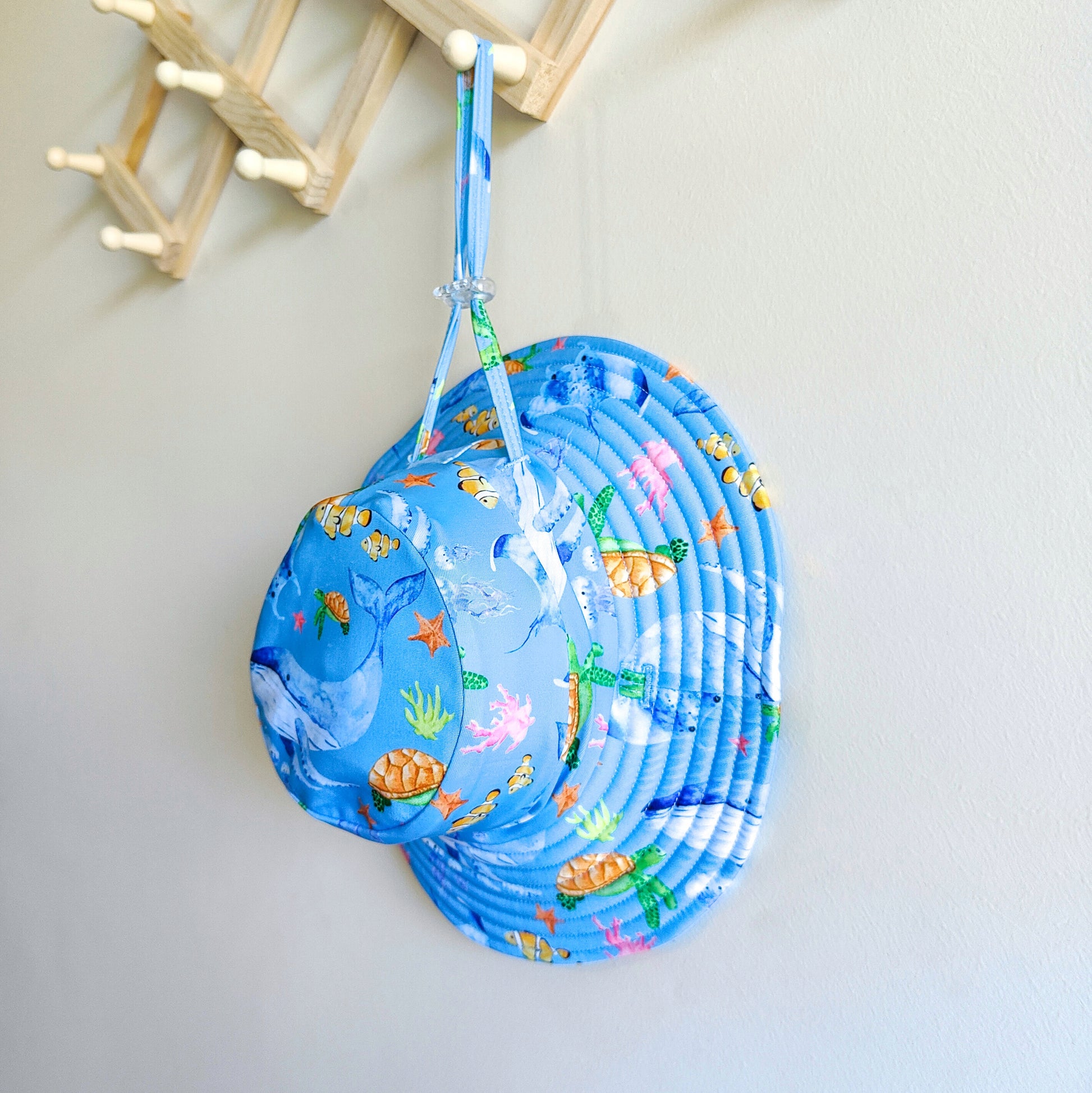 Kids swim hat in blue ocean print with whales, turtles, fish, and coral, made with UPF 50+ sun protection fabric and adjustable toggles for a secure fit.