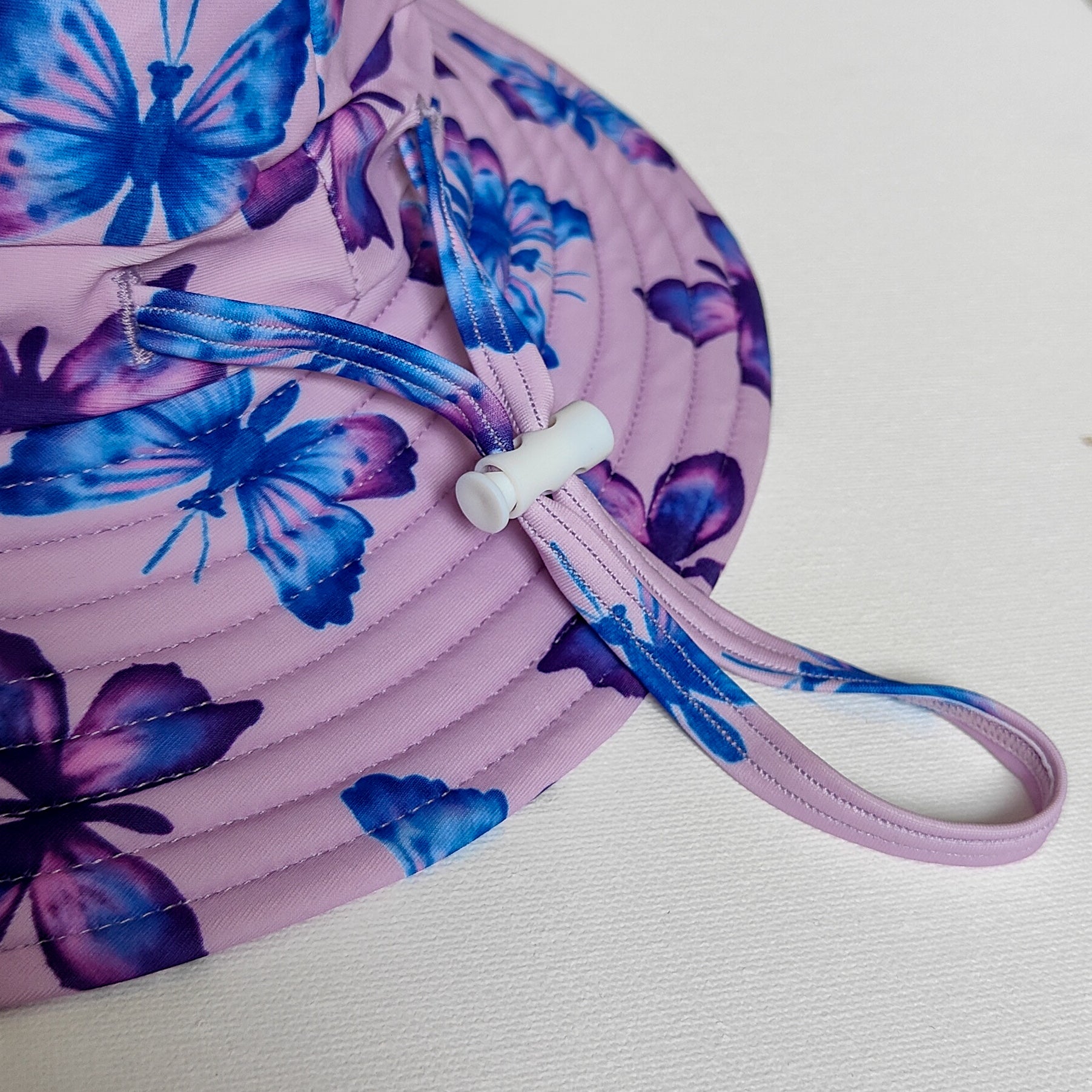 LITTLE E & CO – kids swim hat, a pastel purple with a colourful pink, blue and purple butterflies. Made with UPF 50+ sun protection fabric. The swim hat features adjustable toggles