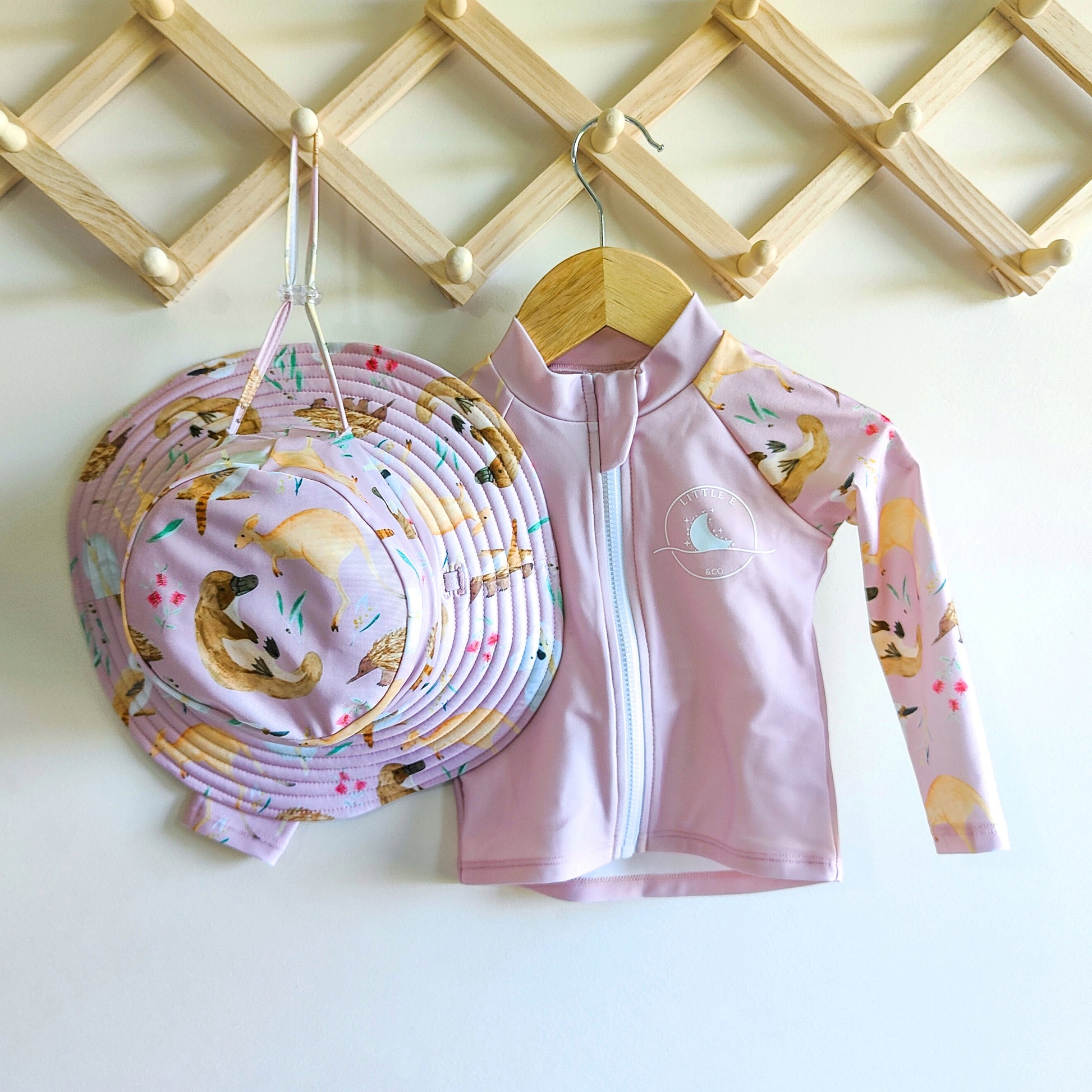 LITTLE E & CO - Two-piece kids' swim set featuring a long-sleeve rash guard and swim hat. The set a pale pink with a colourful Australian animals print featuring koalas, kangaroos, platypus, kookaburras and echidnas. Made with UPF 50+ sun protection fabric. The rash guard has a front zipper for easy wear, and the hat features adjustable toggles.