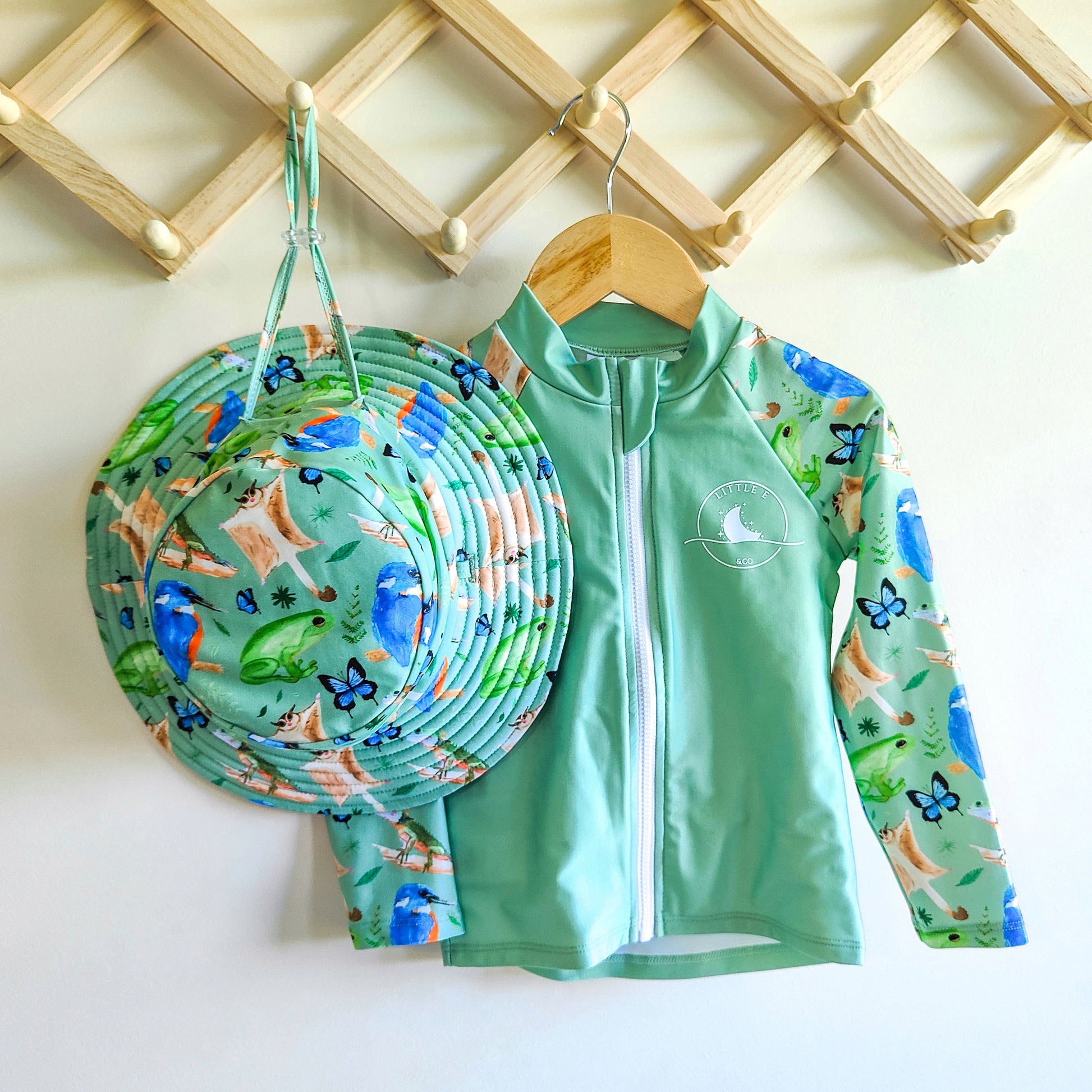 LITTLE E & CO - Two-piece kids' swim set featuring a long-sleeve rash guard and swim hat. The set is a green with a colourful rainforest print featuring birds, frogs, lizards and butterflies. Made with UPF 50+ sun protection fabric. The rash guard has a front zipper for easy wear, and the hat features adjustable toggles.