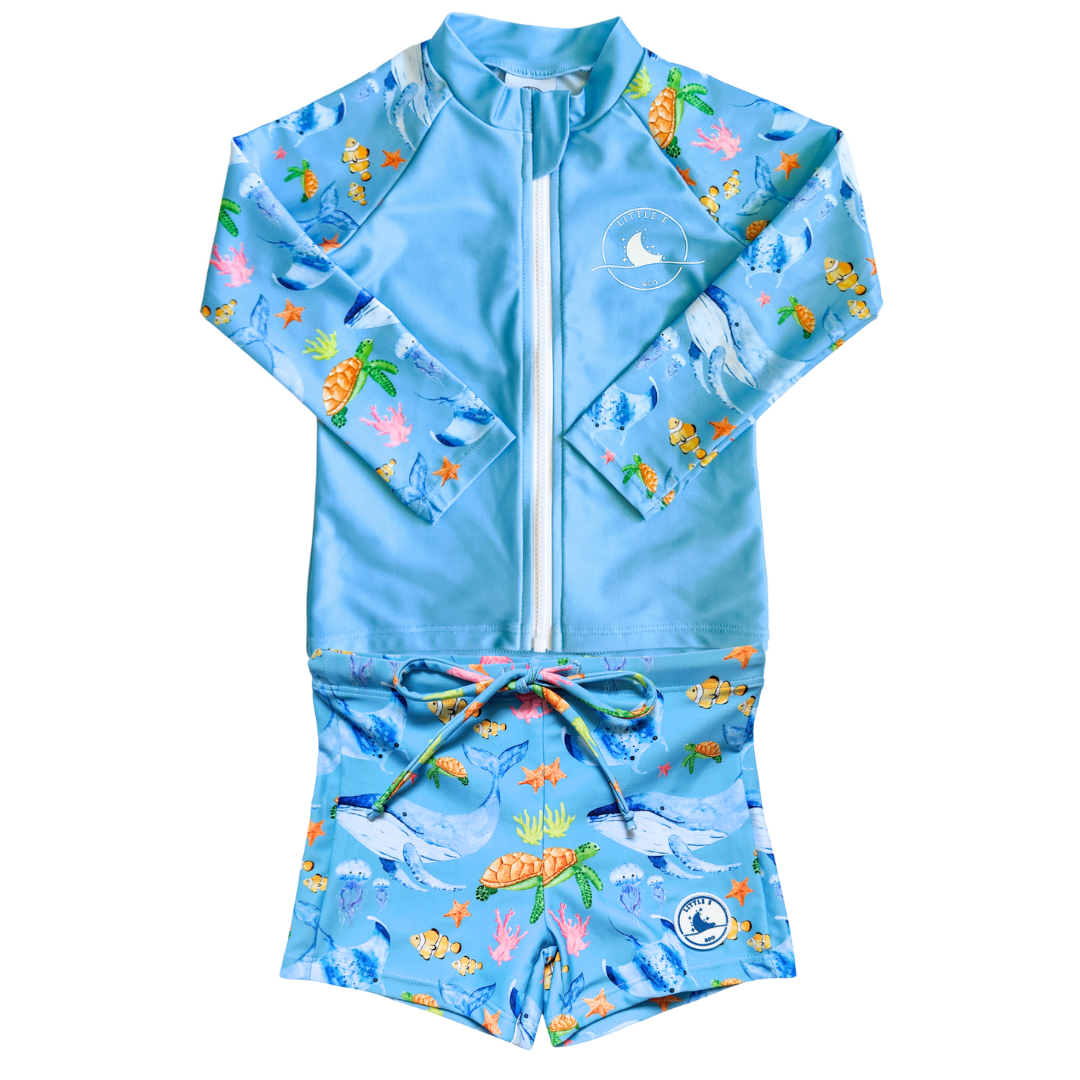 LITTLE E & CO - Two-piece kids' swim set featuring a long-sleeve rash guard and swim shorts. The set is blue with a colourful ocean print featuring whales, turtles, fish and coral. Made with UPF 50+ sun protection fabric. The rash guard has a front zipper for easy wear, and the shorts feature an adjustable drawstring waistband for a secure fit.