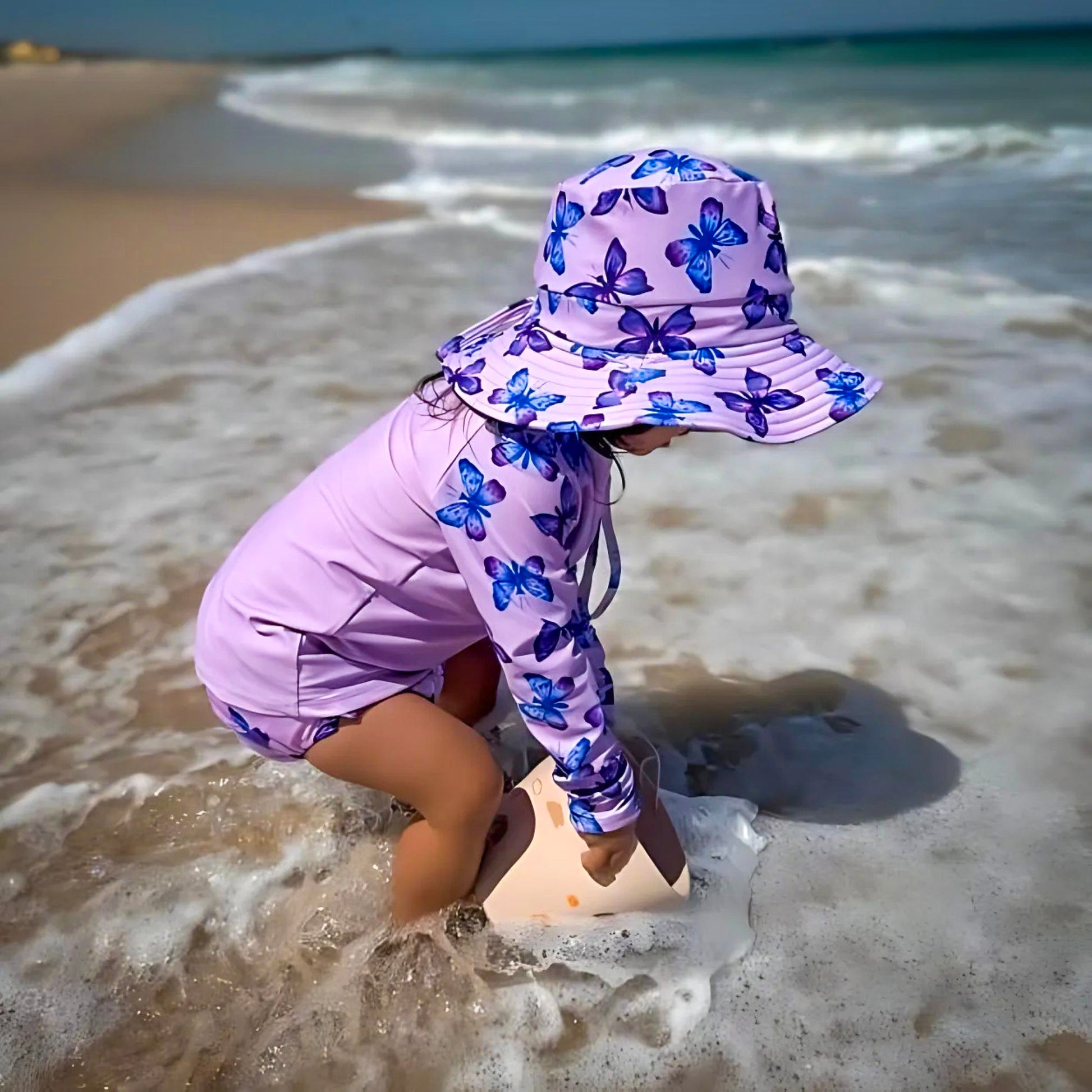 Baby Swimwear Set with pastel butterfly print worn by kid playing at the seaside, featuring long sleeve rashie, swim nappy, and sun hat for sun protection.