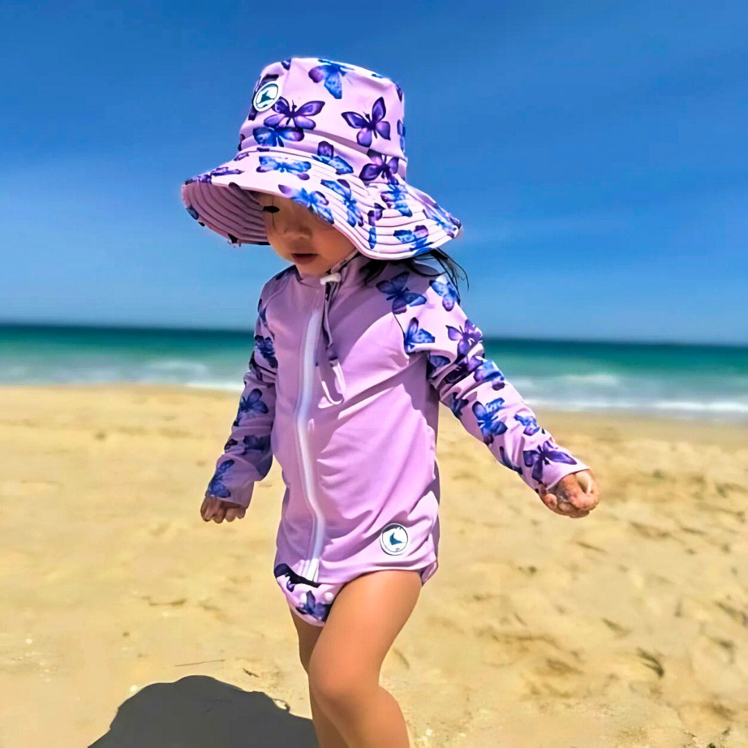 Baby Swimwear Set in pastel butterfly print with UPF 50+ long sleeve rashie, reusable swim nappy, and matching sun hat, worn at the beach.