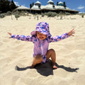 Baby Swimwear Set with butterfly print worn by child playing in sand, featuring long sleeve rashie, swim nappy, sun hat, and wet bag.