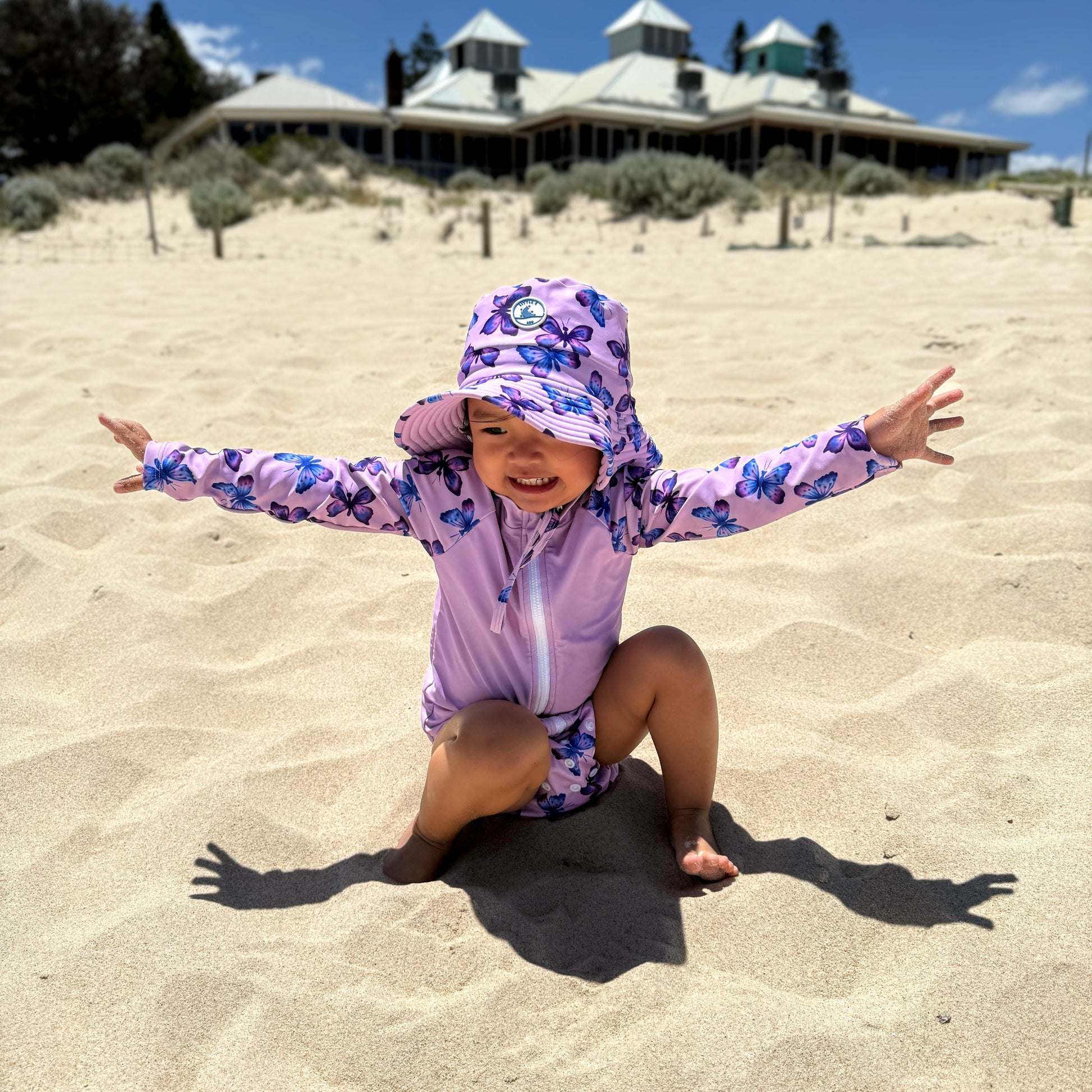 Baby Swimwear Set with butterfly print worn by child playing in sand, featuring long sleeve rashie, swim nappy, sun hat, and wet bag.