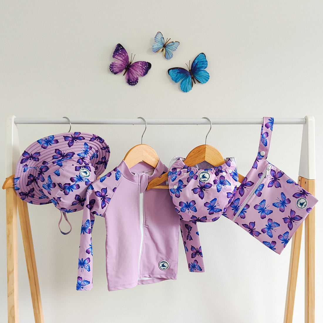 LITTLE E & CO - four-piece kids' swim set featuring a long-sleeve rash guard, swim hat, swim nappy and wet bag. The set a pastel purple with a colourful pink, blue and purple butterflies. Made with UPF 50+ sun protection fabric. The rash guard has a front zipper for easy wear, and the swim nappy has adjustable snaps, and the hat has adjustable toggles.