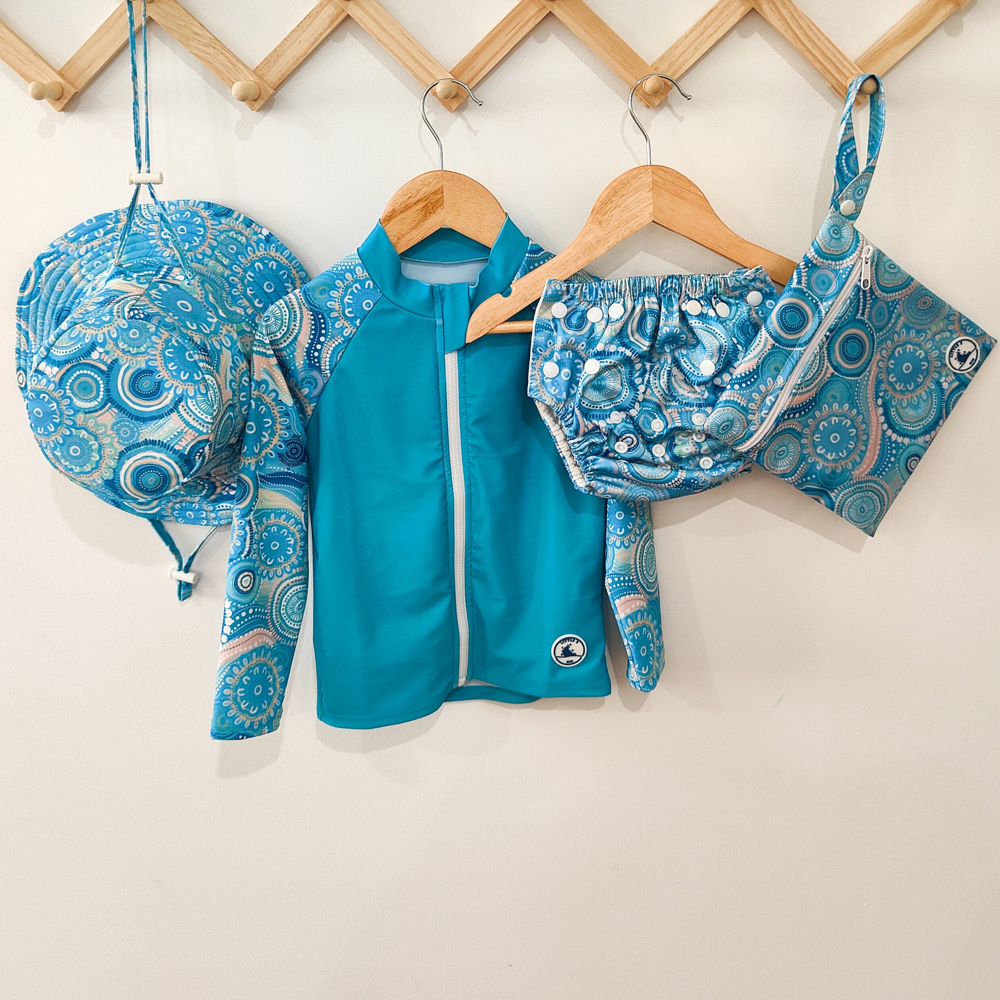 Baby swimwear set in turquoise blue with colourful Indigenous print, includes long-sleeve rashie, adjustable swim nappy, sun hat, and matching wet bag.