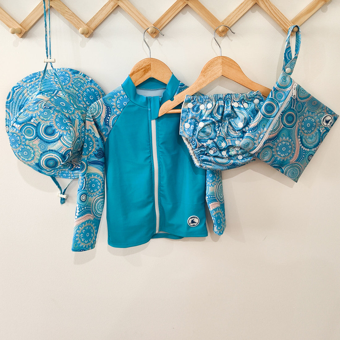 LITTLE E & CO - four-piece kids' swim set featuring a long-sleeve rash guard, swim hat, swim nappy and wet bag. The set is a turquoise blue with a colourful contemporary indigenous print. Made with UPF 50+ sun protection fabric. The rash guard has a front zipper for easy wear, and the swim nappy has adjustable snaps, and the hat has adjustable toggles.
