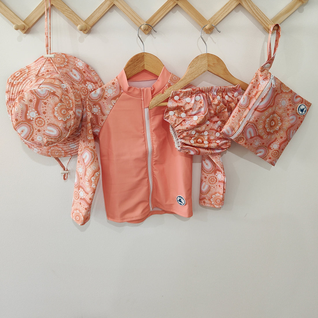 LITTLE E & CO - four-piece kids' swim set featuring a long-sleeve rash guard, swim hat, swim nappy and wet bag. The set is a peach pink with a colourful contemporary indigenous print. Made with UPF 50+ sun protection fabric. The rash guard has a front zipper for easy wear, and the swim nappy has adjustable snaps, and the hat has adjustable toggles.