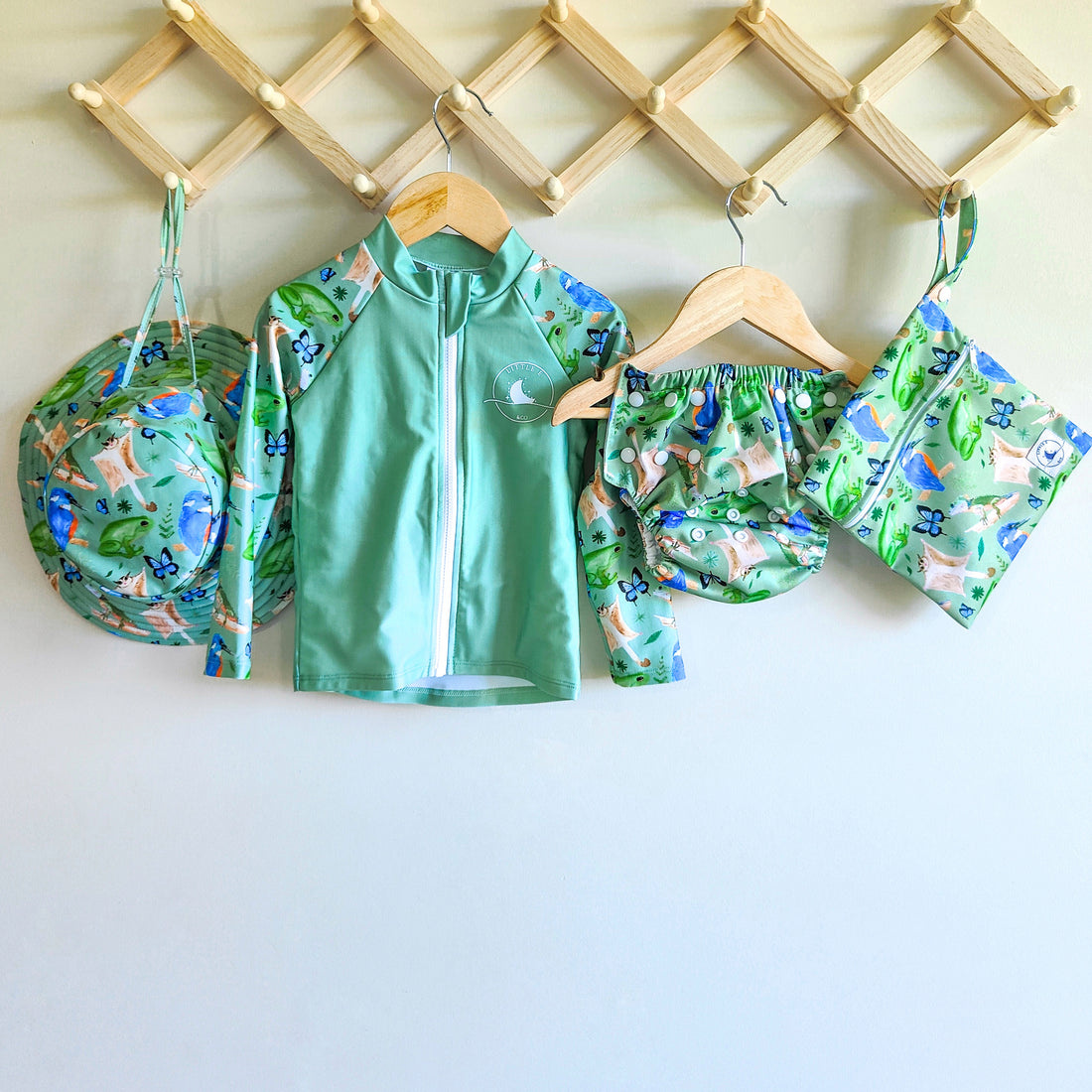 LITTLE E & CO - four-piece kids' swim set featuring a long-sleeve rash guard, swim hat, swim nappy and wet bag. The set is a green with a colourful rainforest print featuring birds, frogs, lizards and butterflies. Made with UPF 50+ sun protection fabric. The rash guard has a front zipper for easy wear, and the swim nappy has adjustable snaps, and the hat has adjustable toggles.