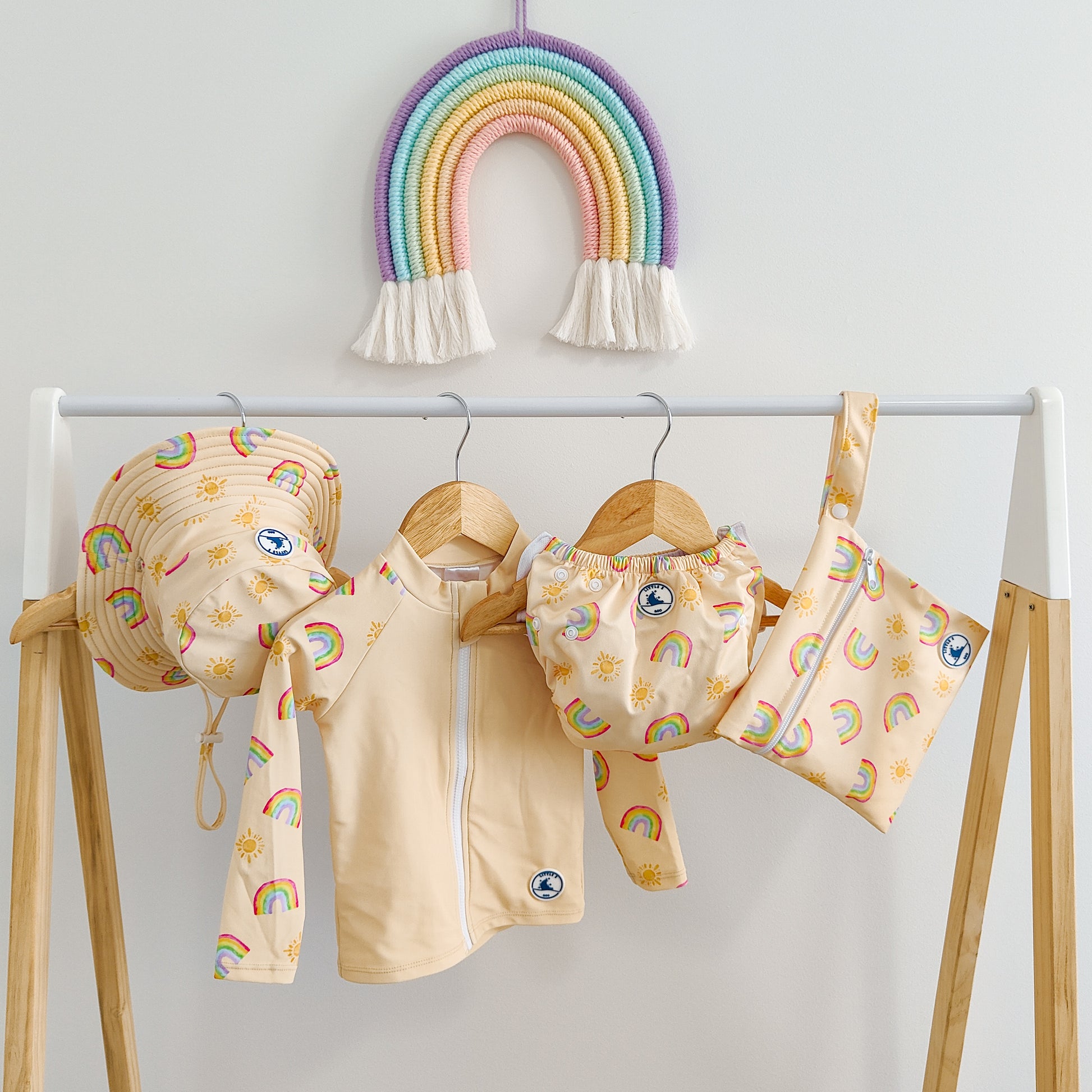 LITTLE E & CO - four-piece kids' swim set featuring a long-sleeve rash guard, swim hat, swim nappy and wet bag. The set a pale yellow with a colourful rainbow and sun print. Made with UPF 50+ sun protection fabric. The rash guard has a front zipper for easy wear, and the swim nappy has adjustable snaps, and the hat has adjustable toggles.