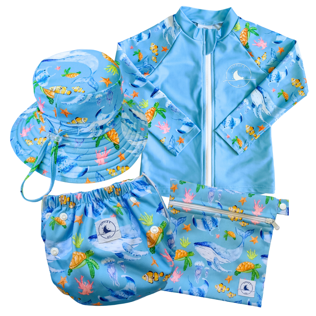 LITTLE E & CO - four-piece kids' swim set featuring a long-sleeve rash guard, swim hat, swim nappy and wet bag. The set is a blue with a colourful ocean print featuring whales, turtles, fish and coral. Made with UPF 50+ sun protection fabric. The rash guard has a front zipper for easy wear, and the swim nappy has adjustable snaps, and the hat has adjustable toggles.