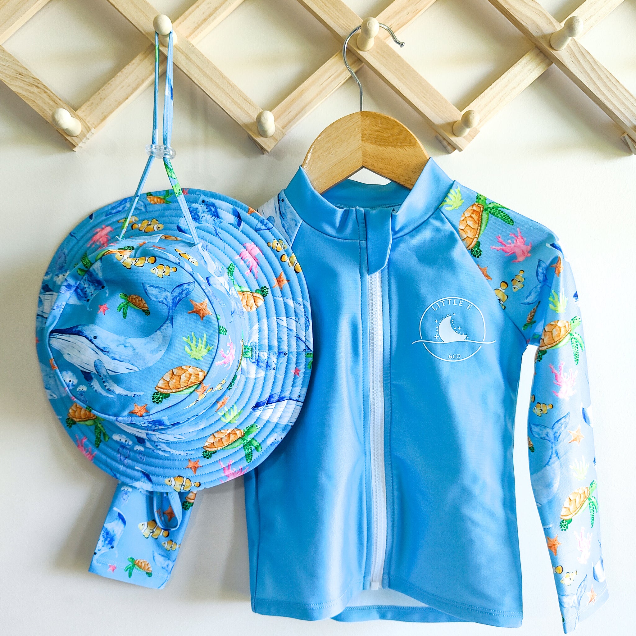 LITTLE E & CO - Two-piece kids' swim set featuring a long-sleeve rash guard and swim hat. The set is a blue with a colourful ocean print featuring whales, turtles, fish and coral. Made with UPF 50+ sun protection fabric. The rash guard has a front zipper for easy wear, and the hat features adjustable toggles.