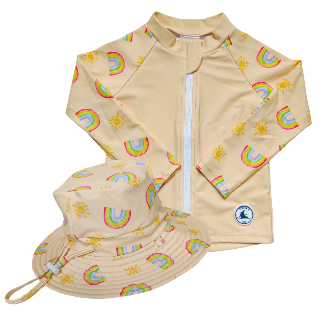 LITTLE E & CO - Two-piece kids' swim set featuring a long-sleeve rash guard and swim hat. The set is a pale yellow with a colourful rainbow and sun print. Made with UPF 50+ sun protection fabric. The rash guard has a front zipper for easy wear, and the hat features adjustable toggles.