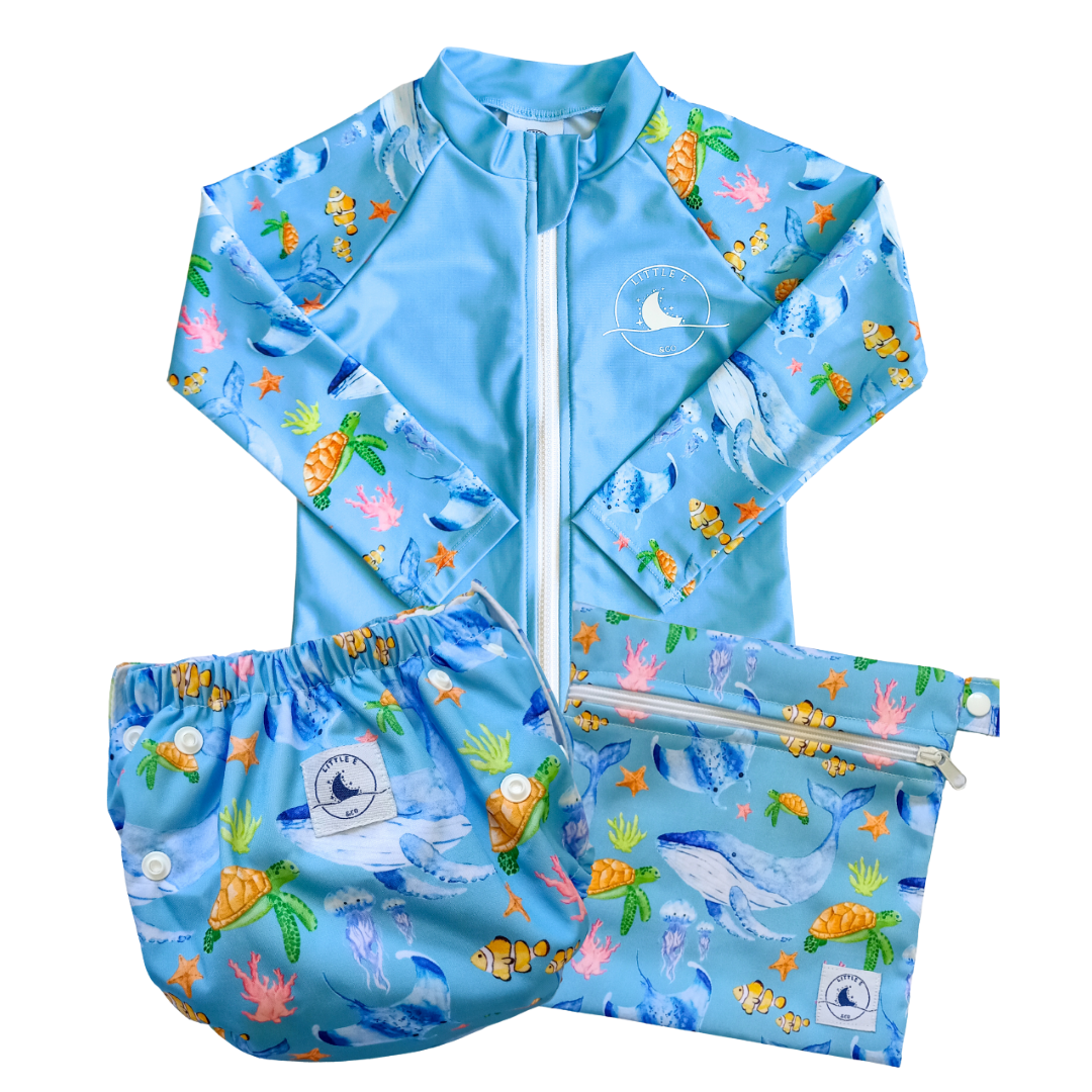 LITTLE E & CO – Three-piece kids' swim set featuring a long-sleeve rash guard, swim nappy and wet bag. The set is a blue with a colourful ocean print featuring whales, turtles, fish and coral. Made with UPF 50+ sun protection fabric. The rash guard has a front zipper for easy wear, the swim nappy has adjustable snaps and the wet bag has a strap.