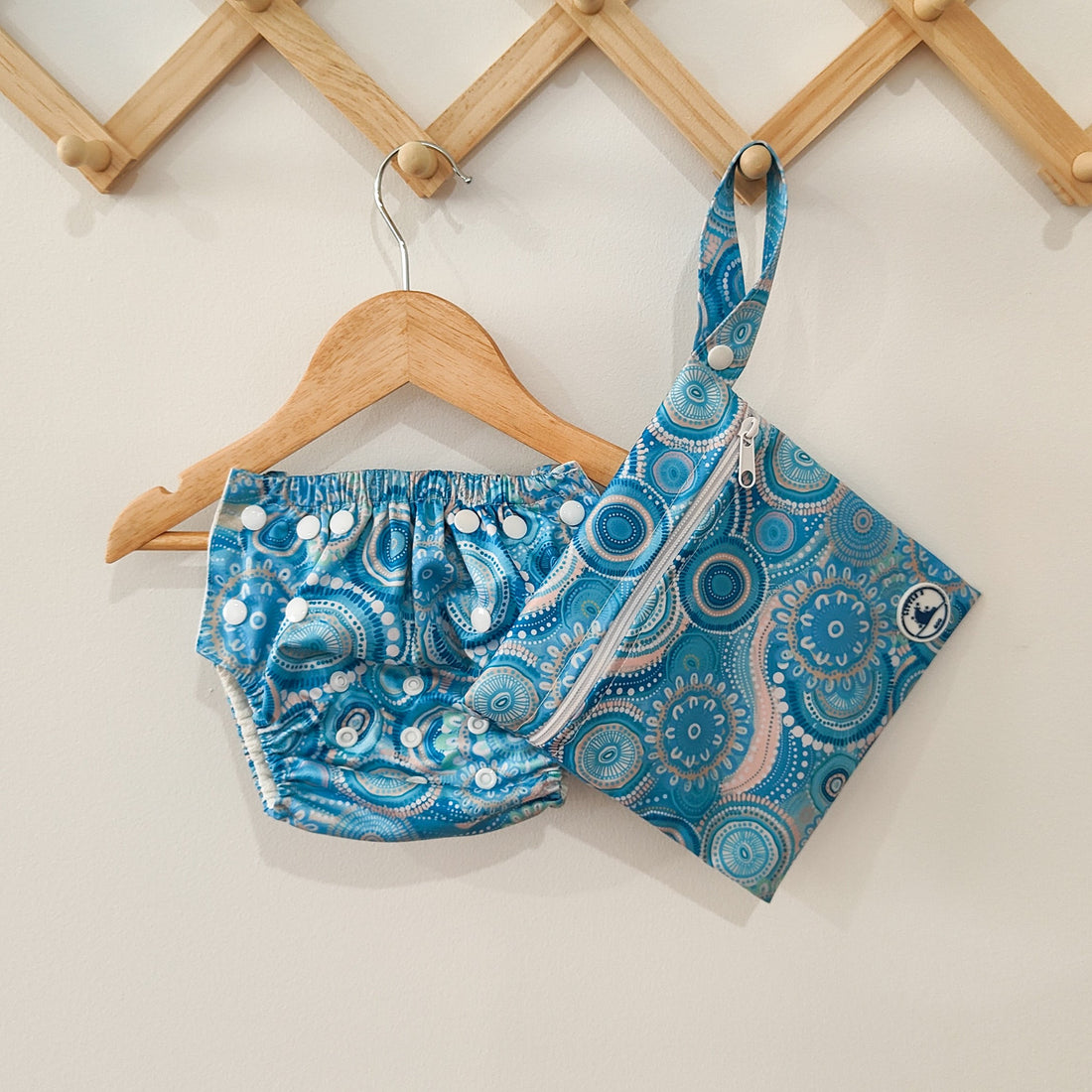 LITTLE E & CO – Three-piece kids' swim set featuring a long-sleeve rash guard, swim nappy and wet bag. The set is a turquoise blue with a colourful contemporary indigenous print. Made with UPF 50+ sun protection fabric. The rash guard has a front zipper for easy wear, the swim nappy has adjustable snaps and the wet bag has a strap.