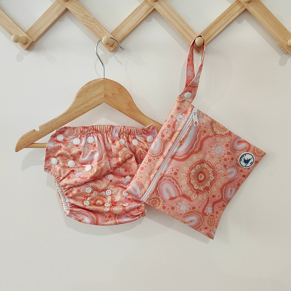 LITTLE E & CO – Three-piece kids' swim set featuring a long-sleeve rash guard, swim nappy and wet bag. The set is a peach pink with a colourful contemporary indigenous print. Made with UPF 50+ sun protection fabric. The rash guard has a front zipper for easy wear, the swim nappy has adjustable snaps and the wet bag has a strap.