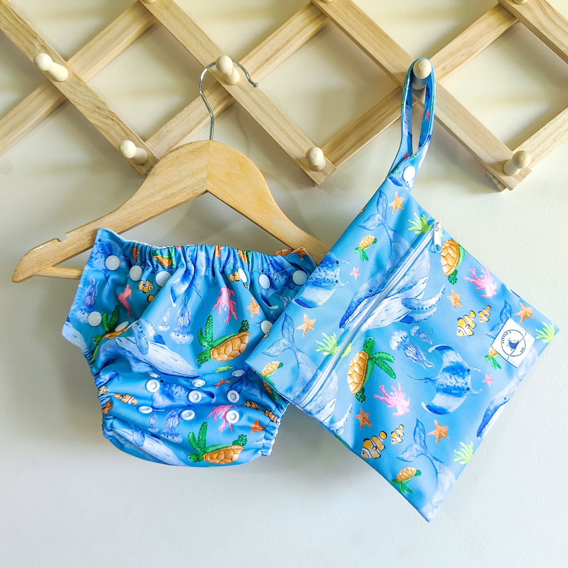 LITTLE E & CO – Two-piece kids' swim set, includes a swim nappy and wet bag, a blue with a colourful ocean print featuring whales, turtles, fish and coral. The swim nappy is a one size fits most design and has 3 adjustable snap settings and the wet bag has a strap.