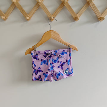 Kids swim shorts in pastel purple with pink, blue, and purple butterfly print, made from UPF 50+ sun protection fabric with adjustable drawstring waistband.

