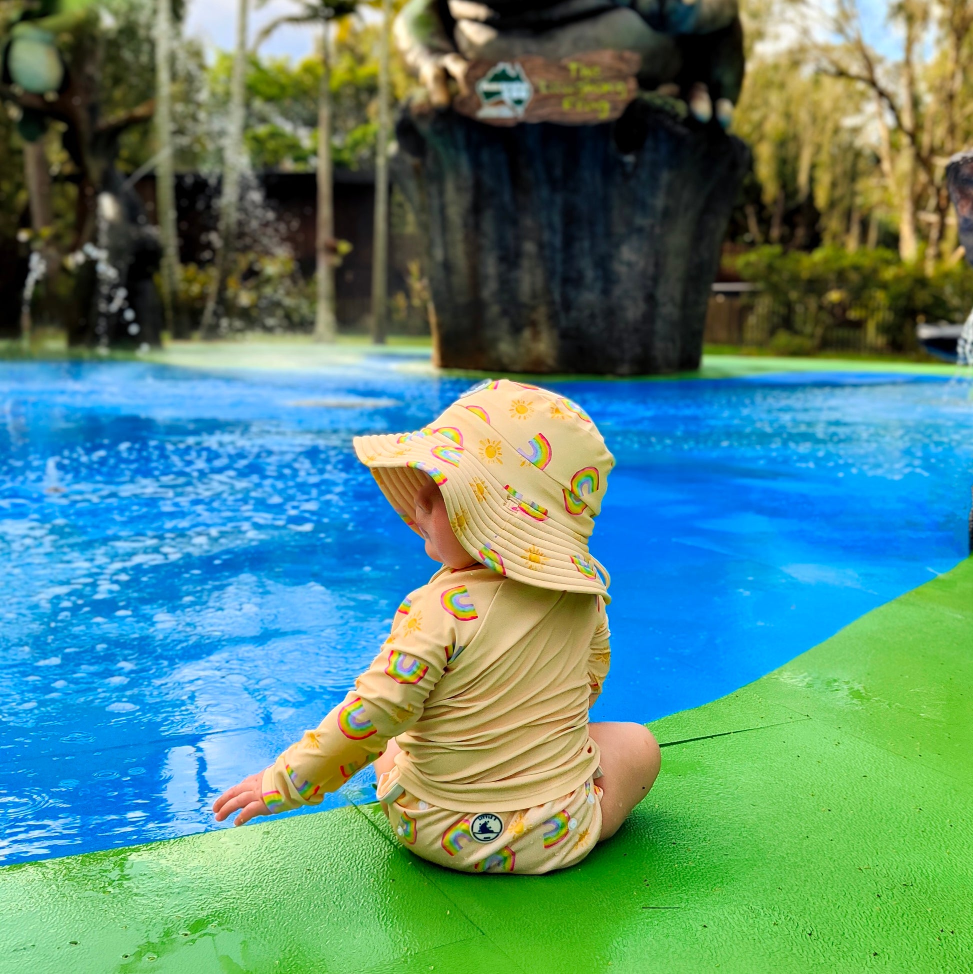 Baby swimwear set worn in pool, featuring pale yellow long sleeve rash guard, swim nappy, and sun hat with rainbow and sun print, made with UPF 50+ protective fabric.

